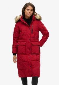 Abrigo acolchado largo rojo con capucha y borde de piel. Cuenta con un bolsillo en el pecho con cremallera, bolsillos laterales y un diseño acolchado con acentos negros.