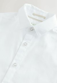 Weißes Hemd aus strukturiertem Stoff mit spitzem Kragen und drei sichtbaren Knöpfen, auf dem die Beschriftung "Baker by Ted Baker" am Knopfleisten zu sehen ist.