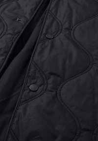 Schwarze gesteppte Jacke mit strukturiertem, wellenförmigem Steppdesign und Druckknöpfen. Das Material ist glatt und leicht.