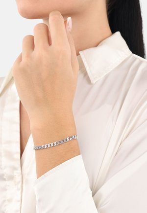 Bracciale in argento con anelli intrecciati, indossato su un polso, su uno sfondo chiaro; abbinato a una camicia bianca con colletto.
