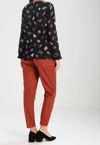 Schwarzes Langarmshirt mit bunten Tier- und Blumenmustern, kombiniert mit taillierten rostfarbenen Hosen und schwarzen Slippern.