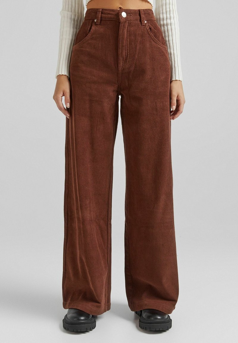 Pantalon en velours côtelé marron taille haute à jambes larges porté avec des chaussures noires épaisses et un pull court côtelé crème.