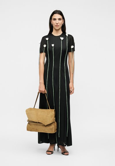 Femme portant une longue robe noire avec des lignes verticales vertes et des détails de fleurs blanches, tenant un grand sac à main en daim beige, portant des talons noirs.