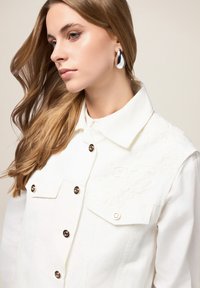 Witte denim jas met zichtbare knoopsluiting, geborduurd bloemenmotief op één schouder en een klassieke kraag. Gladde textuur, casual ontwerp.