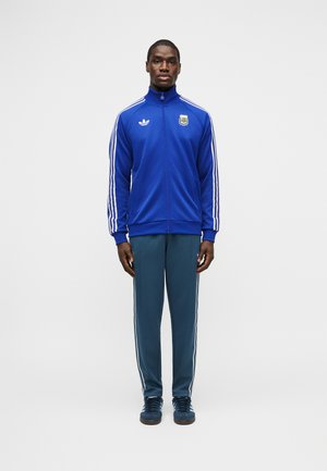 Uomo in piedi, volto rivolto in avanti, che indossa una giacca sportiva Adidas blu con strisce bianche e logo dell'Associazione di Calcio Argentina, pantaloni tuta blu e scarpe da ginnastica blu.
