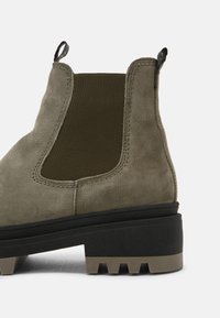 Botín Chelsea de ante en verde oliva con paneles laterales elásticos, acabado texturizado y suela de goma negra gruesa con detalles de tracción.
