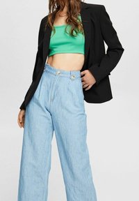 Femme aux longs cheveux vêtue d'un blazer noir, d'un top court vert et d'un jean large taille haute bleu clair sur un fond uni.
