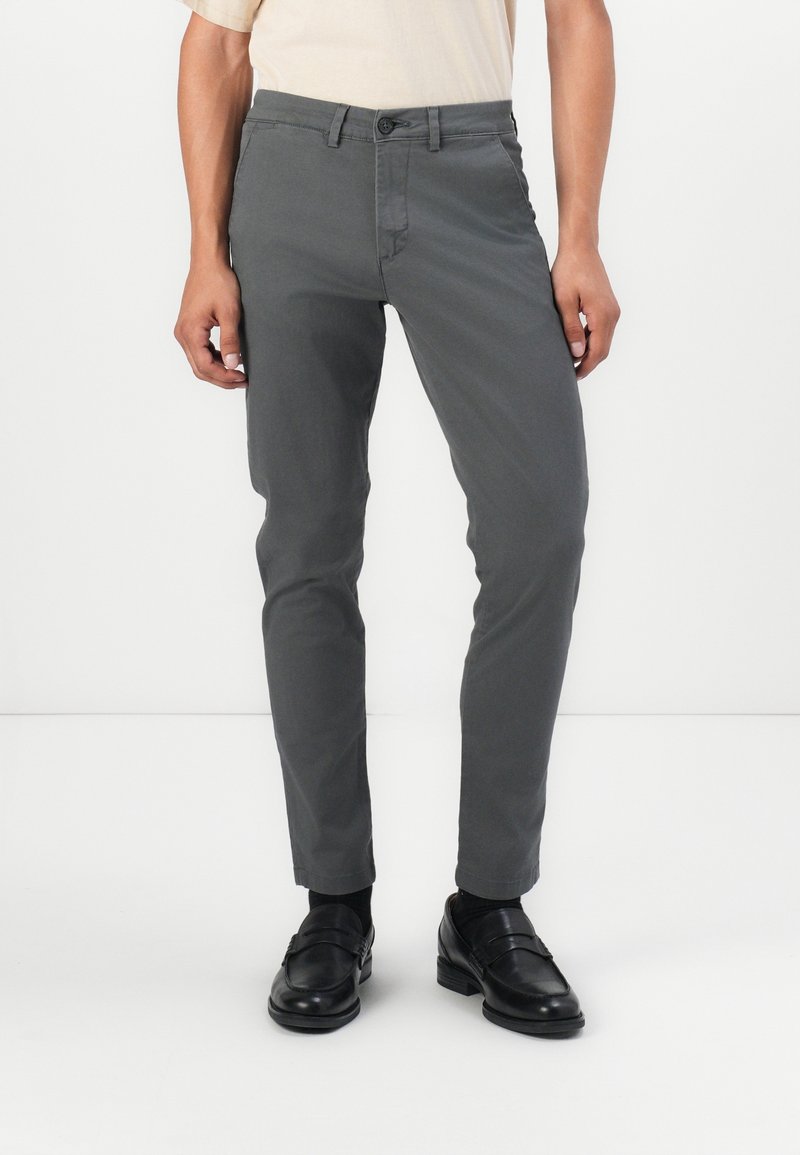 Pantalon chino gris au style slim, doté d'une fermeture par bouton et fermeture éclair, poches latérales, et tissu en mélange de coton lisse. Associé à des mocassins noirs.