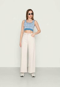 Mulher jovem a usar óculos de sol, top curto azul com padrão floral, calças brancas de perna larga de cintura alta e sapatilhas brancas, de pé com uma mão no bolso.