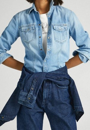 Person iført en lyseblå denimskjorte over en hvid grafisk t-shirt, med en mørkeblå denimjakke med prikker bundet om livet og matchende bukser.