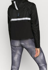 Czarna lekka kurtka z kapturem, z białym poziomym paskiem i nadrukowanym tekstem. W połączeniu z dopasowanymi, wzorzystymi szarymi legginsami.