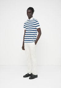 Άνδρας που στέκεται με τα χέρια στις τσέπες, φοράει λευκό παντελόνι, μαύρα μοκασίνια και μπλε, μαύρο και λευκό ριγέ πόλο πουκάμισο.