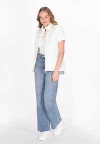 Vit vadderad väst med hög krage, bärs över en vit skjorta, kombinerad med ljusblå vida jeans och vita sneakers.