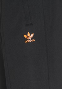 Svarta sweatpants i mjukt tyg har en orange Adidas-logotyp med en trebladig design, placerad ovanför fållen på vänster ben.