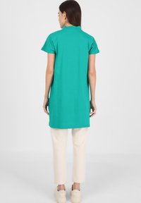 Camicia da uomo turchese con colletto, maniche corte, in tessuto morbido, lunghezza al ginocchio, abbinata a pantaloni color crema e calzature chiare.