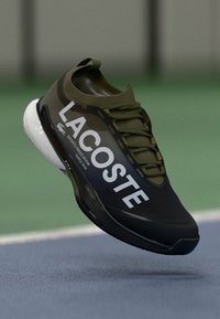 Lacoste atletska cipela u tamno maslinastoj boji s elegantnim, glatkim dizajnom. Ima bijeli logo, zaobljen vrh i reljefne vezice.