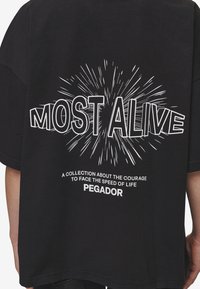 Zwart katoenen T-shirt met witte grafische tekst "MOST ALIVE" met uitstralende lijnen en de merknaam "PEGADOR" eronder.
