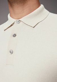 Camisa polo beige hecha de tejido de punto, con cuello tipo polo y tres botones metálicos, que muestra una textura suave y un diseño ajustado.