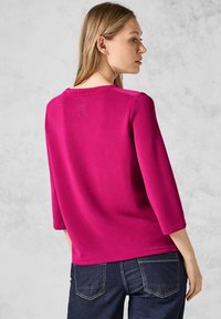 Un haut magenta à manches longues en tissu lisse, avec un col rond et un petit détail graphique au dos. Associé à un jean noir.