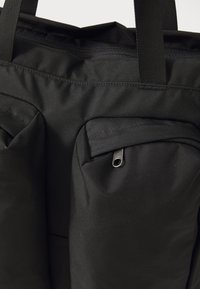 Zbliżenie czarnej torby na ramię z dwoma przednimi kieszeniami na zamek błyskawiczny i mocnymi, plecionymi uchwytami, wykonanej z wytrzymałego materiału.