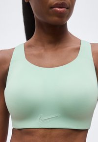 Žena odjevena u svijetlozelenu Nike sportsku majicu, skračenu na području struka i vrata, na jednostavnoj svijetloj pozadini.