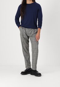 Pull-over à col rond bleu marine, pantalon à carreaux gris avec plis et chaussures noires. Le pantalon présente un motif texturé dans des teintes plus claires.