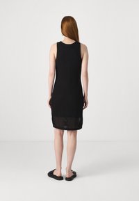 Vestido preto sem mangas apresentando um padrão texturizado semelhante a uma malha. A barra é ligeiramente transparente, combinado com sapatos pretos simples que se calçam facilmente.