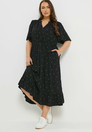 Frau trägt ein schwarzes Midikleid mit weißen Punkten, flatternden Ärmeln, Taschen und gerafftem Saum, kombiniert mit weißen Sneakers, steht an einer Wand.