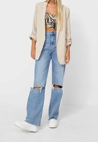 Beige oversized blazer gedragen over een patroonrijke cropped top, gecombineerd met lichtblauwe gescheurde wijde jeans en witte sneakers.