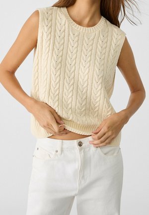 Pull sans manches crème en maille torsadée porté par une personne ajustant l'ourlet, associé à un jean blanc taille haute avec fermeture à boutons.
