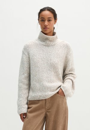 Vrouw met donker haar die een losse lichtgrijze coltrui en bruine ribfluwelen broek draagt, staand met één hand in de zak.