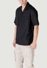 Camisa negra de manga corta hecha de una tela suave, con dos bolsillos en el pecho y un diseño de cuello tipo camp. Sencilla, con cierres de botones.