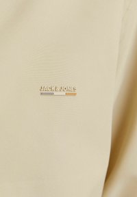 Țesătură bej cu o textură netedă, având un mic logo auriu „JACK & JONES” și trei dungi colorate (gri, alb, portocaliu) dedesubt.