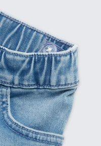 Jasnoniebieski pas do spodni dżinsowych z regulowaną gumką od środka i małym przejrzystym guzikiem na jasnym tle.