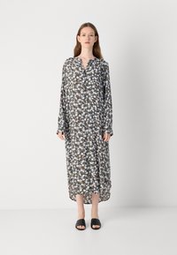 Robe lunga con bottoni e motivo floreale in tonalità di verde, bianco e blu. Il tessuto è leggero e semi-trasparente. Sandali neri da infilare.