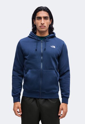 Homme à la peau moyenne et aux cheveux noirs portant un sweat à capuche zippé bleu marine The North Face et un pantalon noir, debout devant un fond uni.