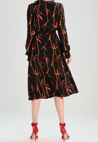 Schwarzes Kleid mit floralem Muster, das rote und orange Akzente aufweist. Lange Ärmel, tailliert und mit fließendem Saum. Rote High-Heels.