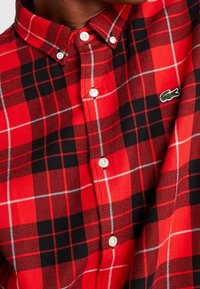 Camisa de cuadros rojos y negros hecha de tejido entrelazado, con un cuello de botonadura, botones blancos y un pequeño logo verde en el pecho.