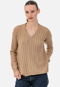 Beiger Pullover mit Zopfmuster, V-Ausschnitt, Rippenstruktur, langen Ärmeln und lockerem Sitz. Der Stoff wirkt weich und warm.