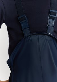 Tumši zili ūdensnecaurlaidīgi kombinezoni ar regulējamiem siksnām, kas raksturojas ar gludu tekstūru un pastiprinātiem šuvēm piestiprināšanas punktos.