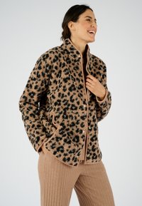 Jasje met luipaardprint in zachte, pluche stof met een tan basis en zwarte vlekken, voorzien van drukknoopjes en zijzakken. Gecombineerd met geribbelde broek.