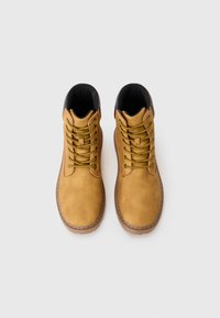 Botas de ante marrón con forro negro, con cordones amarillos y un diseño de punta redonda. Observa los detalles de costura a lo largo de la suela.