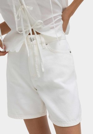 Hvide højtaljede denimshorts med lommer foran og bagpå, båret af en person i en hvid top med snørelukning og bølget kant.