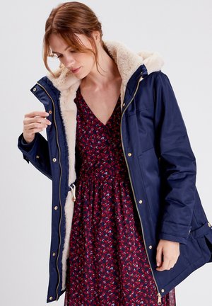 Parka marine avec quincaillerie dorée, dotée d'une douce doublure en polaire crème et d'une capuche. Portée par-dessus une robe florale rouge avec un décolleté en V.