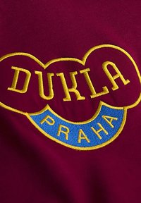 Bordeaux textiel met gele geborduurde tekst "DUKLA" in hartvorm, met daaronder een blauwe band waarop het woord "PRAHA" staat.