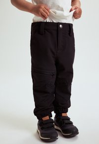 Kind met zwarte cargobroeken met elastische manchetten en klittenbandschoenen, die een wit shirt recht trekt, tegen een effen lichte achtergrond.