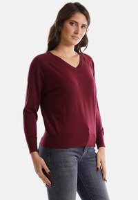 Donna con capelli lunghi castani che indossa un maglione bordeaux a scollo a V e jeans blu, in posa con le mani vicino ai fianchi su sfondo bianco.