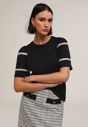 Maglietta nera a maniche corte con dettagli bianchi simili a pizzo sulle maniche, abbinata a una gonna nera e bianca a motivi testurizzati con strisce orizzontali.