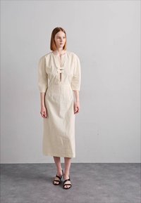 Rochie bej, până la genunchi, cu mâneci bufante și închidere frontală în formă de cheie, confecționată dintr-un material texturat. Purtați-o cu sandale negre cu sireturi.
