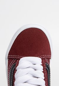 Baskets en daim rouge avec lacets blancs et semelle en caoutchouc, présentant des bandes latérales noires et des coutures contrastées le long des bords.